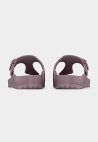 Ζευγάρι μοβ σανδαλιών slip-on με υφασμένη σόλα και διπλές λουρίδες με αγκράφες, που φαίνεται από πίσω σε απλό ανοιχτό φόντο.