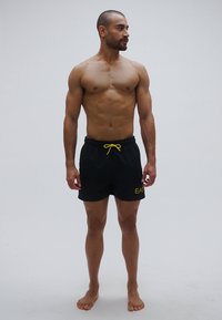 Fekete fürdőnadrág sárga húzózsinórral és logóval. Könnyű anyagból készült, laza szabással, térd feletti fazonban.
