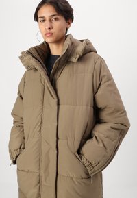 Parka marron avec capuche, dotée d'un design matelassé, de poignets élastiques et de poches latérales. Fabriquée en tissu doux et texturé.