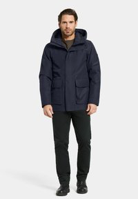 Marine hoodie gemaakt van waterafstotend materiaal; beschikt over twee voorzakken en een ritssluiting; gecombineerd met zwarte broeken en laarzen.