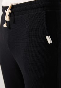Schwarze, strukturierte Jogginghosen mit einem Kordelzugbund, seitlichen Taschen und einem sichtbaren Etikett auf der linken Tasche. Aus einem weichen, gerippten Stoff gefertigt.