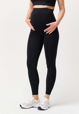 Pregnant woman wearing black maternity leggings and white athletic shoes, hands gently resting on belly.