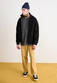 Sort strikket zip-jakke over en grå skjorte, parret med tan brede bukser og sorte sneakers med hvide såler. Marineblå hue på hovedet.