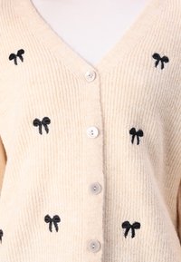 Gilet crème avec texture en côtes verticales, présentant des détails en forme de nœud noir brodés et quatre boutons blancs à l'avant.
