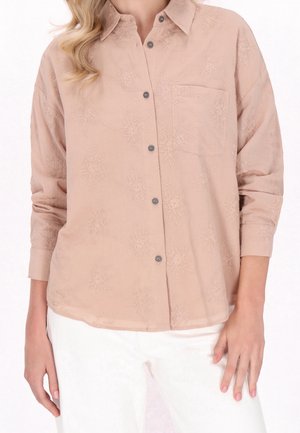 Femme portant un chemisier beige à boutons avec broderie florale et pantalon blanc, debout les bras détendus sur les côtés.