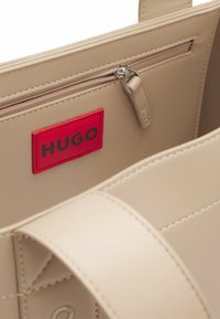 Beige käsilaukku, jossa on sileä, synteettinen pinta. Varustettu punaisella HUGO-logo-patchilla ja sisäisellä vetoketjullisella taskulla. Pyöreät hihnat.