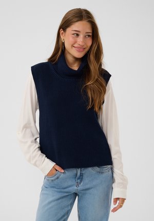 Junge Frau mit langen Haaren, die goldene Ohrringe trägt, einen marineblauen ärmellosen Rollkragenpullover über einem weißen Langarmhemd und hellblaue Jeans.