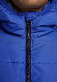 Niebieska pikowana kurtka z wysokim kołnierzem, zamkiem błyskawicznym z przodu oraz gładką teksturą. Charakterystyczny wzór przeszyć na całym materiale.