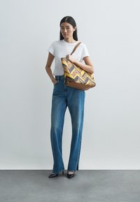 Vícebarevná taška přes rameno s geometrickým vzorem v žlutých a hnědých tónech a širokým popruhem. Modelka má na sobě bílou košili a modré džíny.