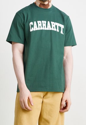Persona que lleva una camiseta verde Carhartt con texto de logo blanco y pantalones marrón claro, de pie contra un fondo llano y claro.