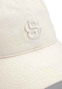 Cappellino da baseball bianco in cotone con logo ricamato in beige chiaro. Presenta una visiera curvata e dettagli cuciti.