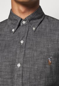 Camisa de botones gris con un tejido texturizado, que presenta botones blancos y un logo bordado de un jugador de polo en el lado izquierdo del pecho.