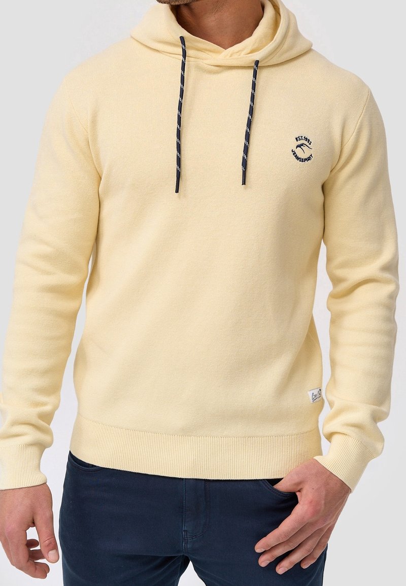 Homme portant un sweat à capuche jaune clair avec des cordons foncés et un petit logo noir sur la poitrine gauche, associé à un pantalon bleu foncé.