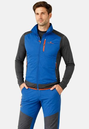 Uomo che indossa un gilet outdoor blu e grigio con dettagli della cerniera arancione sopra una maglia nera a maniche lunghe, in piedi con le mani nelle tasche.