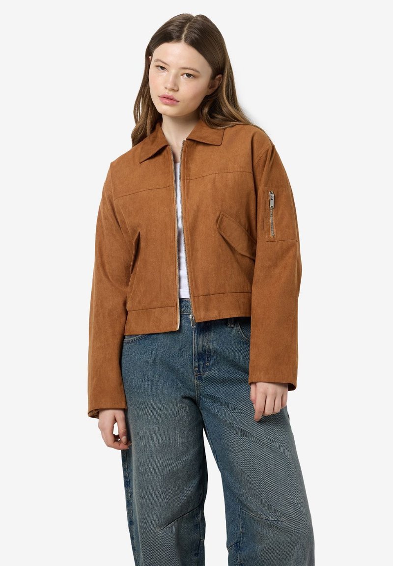 Veste en suédine marron, courte, avec un col, fermeture éclair sur le devant et poches latérales. Comprend une poche zippée sur le bras.