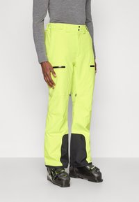 Άτομο που φοράει φωσφορούσα κίτρινη σκιέρ παντελόνι με μαύρα ενισχυμένα μπατζάκια και μαύρες σκιές μπότες, στέκεται μπροστά σε απλό φόντο.