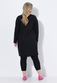 Robe noire à manches longues avec taille froncée, associée à des leggings noirs et des chaussures à enfiler roses. Tissu lisse et design à hauteur du genou.
