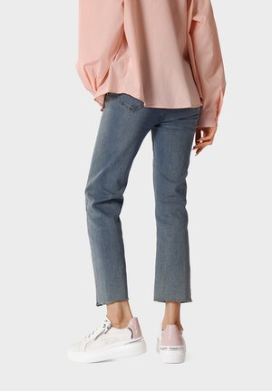 Donna che indossa jeans blu corti, sneakers bianche con zeppa e tacco rosa, e una maglietta rosa chiaro a maniche lunghe e larghe, in piedi su sfondo bianco.