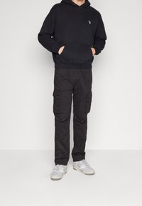 Person som bär en svart huvtröja, svarta cargobyxor med sidofickor och vita sneakers, står med händerna i huvtröjans ficka.