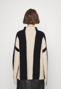 Personne aux cheveux bruns courts portant un pull douillet beige à rayures noires, vue de dos, associée à une jupe en cuir noire.