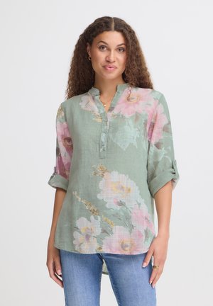 Kvinde med krøllet hår iført en lysegrøn blomstret bluse med opkæmpede ærmer og blå jeans, stående mod en ensfarvet baggrund.