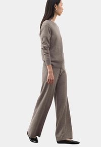 Donna che cammina indossando un maglione lavorato a maglia color taupe e pantaloni a gamba larga con ballerine nere su uno sfondo chiaro e semplice.