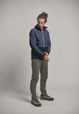 Mann richtet den Ärmelbund einer farbblockierten Jacke mit navyblauen und schwarzen Bereichen, trägt graue Hosen und Wanderschuhe, steht vor einfachem Hintergrund.