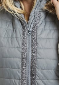 Graue Pufferjacke mit einem dekorativen silbernen Reißverschluss und strukturiertem Einsatz in der Mitte, mit einer Kapuze, die mit Kunstpelz gefüttert ist.