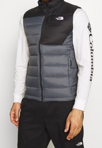 Gesteppte Weste in Grau mit einem schwarzen oberen Abschnitt, ausgestattet mit hohem Kragen, Reißverschluss und Seitentaschen. Hergestellt aus leichtem, gestepptem Material.