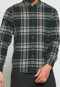 Groen en grijs geruite button-up overhemd van flanel, met een puntige kraag, lange mouwen en zwarte knopen aan de voorkant.