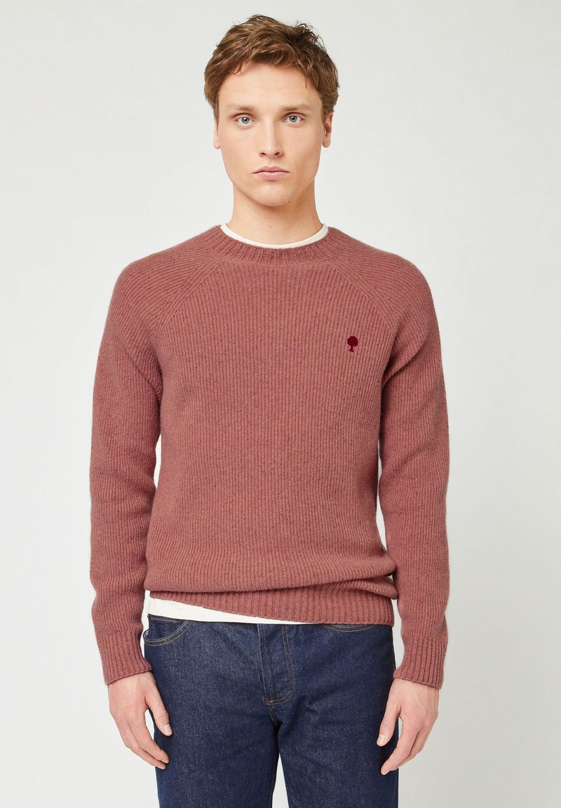 Junger Mann, der einen gerippten mauvefarbenen Pullover mit einem kleinen Baum-Logo und dunkelblauen Jeans trägt, vor einem schlichten hellen Hintergrund steht.