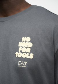 Κοντινό πλάνο γκρι πουκαμίσου με κειμήλιο σε εκρού γράμματα που αναγράφει «NO NEED FOR TOOLS» και το λογότυπο «EA7 Emporio Armani» κάτω από το κείμενο στο στήθος.