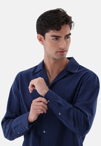 Camicia blu navy con colletto a punta, tessuto testurizzato e maniche lunghe. Presenta chiusure a bottone visibili sui polsini.