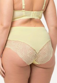 Lace-geborduurde, hooggewaade slips in pastelgeel met een gladde voorkant en intricaten kantdetails langs de achterranden.