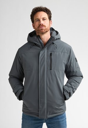 Mann trägt graue Kapuzen-Winterjacke mit vorne geschlossenem Reißverschluss, Hände in den Taschen, steht vor schlichtem hellem Hintergrund.