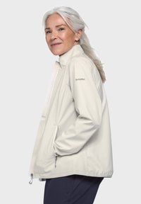 Helle beige Softshell-Jacke mit hohem Kragen, seitlichen Taschen und einem Logo am Ärmel. Der Stoff sieht glatt aus und hat einen leichten Glanz.