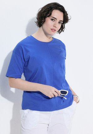 Kobieta z krótkimi ciemnymi włosami, ubrana w niebieski t-shirt i białe spodnie, trzymająca okulary przeciwsłoneczne z białą oprawą na jednolitym białym tle.