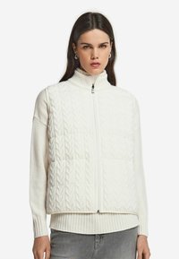 Kvinna som bär en vit kabelstickad ärmlös zip-up väst över en vit ribbad tröja med grå byxor, stående mot en vit bakgrund.