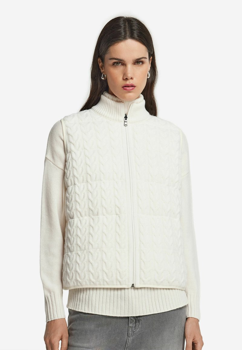 Kvinna som bär en vit kabelstickad ärmlös zip-up väst över en vit ribbad tröja med grå byxor, stående mot en vit bakgrund.