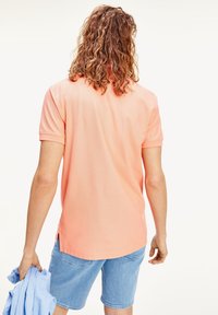 Kortärmad polo i ljus korall, tillverkad av texturerad tyg, med en subtil sömslits; person som håller en ljusblå skjorta i vänster hand.