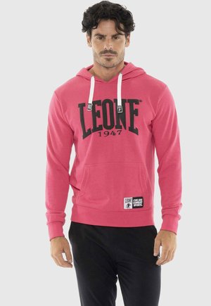 Mann steht und trägt einen pinkfarbenen LEONE 1947 Hoodie mit weißen Kordeln und schwarzen Hosen vor einem einfarbigen Hintergrund.