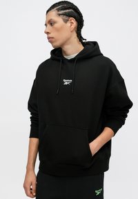 Felpa nera con cappuccio realizzata in misto cotone, con tasca a marsupio frontale e logo Reebok ricamato in bianco. Vestibilità ampia, polsini a costine.