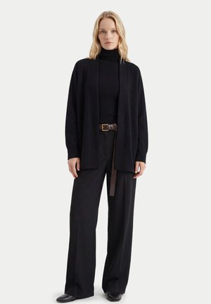 Frau mit blonden Haaren, die einen schwarzen Rollkragenpullover, eine offene Strickjacke, weite Hosen, einen braunen Gürtel und schwarze Schuhe trägt, steht vor weißem Hintergrund.