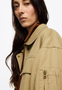 Chaqueta caqui con un cuello marrón, que cuenta con cierre de cremallera, bolsillos frontales y una tela texturizada. Cabello largo y oscuro cae sobre el hombro.
