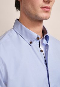 Hellblaues Hemd mit strukturiertem Kragen, braunen Knopfakzenten und einer strukturierten Stoffoberfläche. Verfügt über eine kontrastierende innere Leiste.