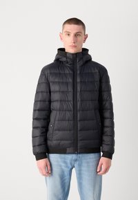 Sort pufferjakke med hætte og lynlås foran, med et kviltet mønster, elastiske manchetter og sidelommer. Båret over lyseblå jeans.