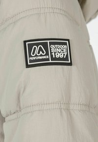 Gai pelēks pūkains jaka ar izšuvumu. Uz piedurknes ir melns gumijas logotipa uzlīme ar tekstu "OUTDOOR SINCE 1997".