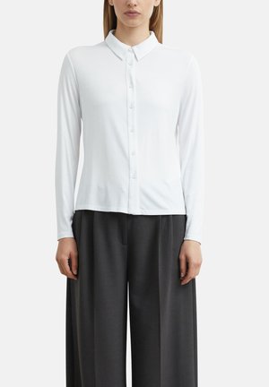Frau trägt eine eng anliegende weiße Hemdbluse mit langen Ärmeln und lockere schwarze weite Hosen, steht vor einem einfarbigen Hintergrund.