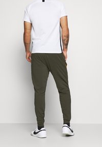 Vit kortärmad skjorta med svarta detaljer och olivgröna joggers, med sidofickor och en avslappnad passform. Fötterna pryds av vita och svarta sneakers.