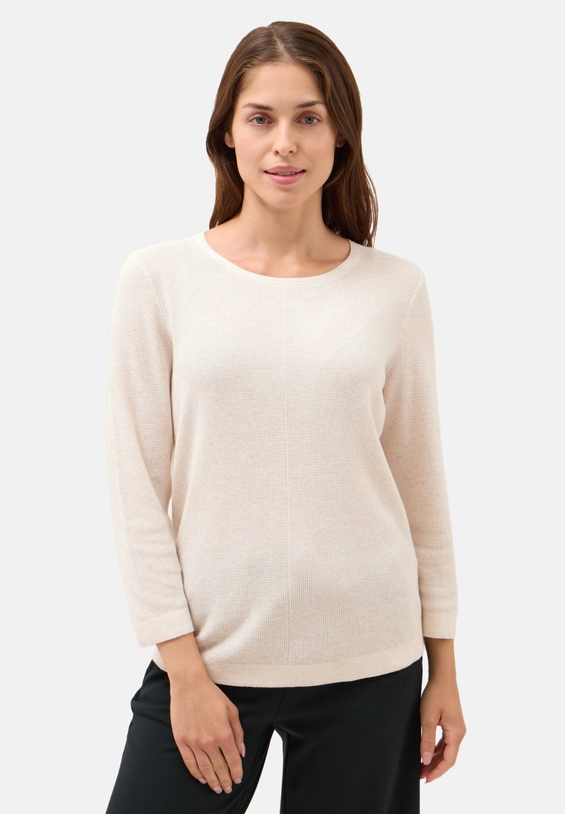 Hellbeige, langärmliger Strickpullover mit Rundhalsausschnitt und strukturiertem Muster, kombiniert mit schwarzen Hosen. Figurbetonte, aber lässige Silhouette.